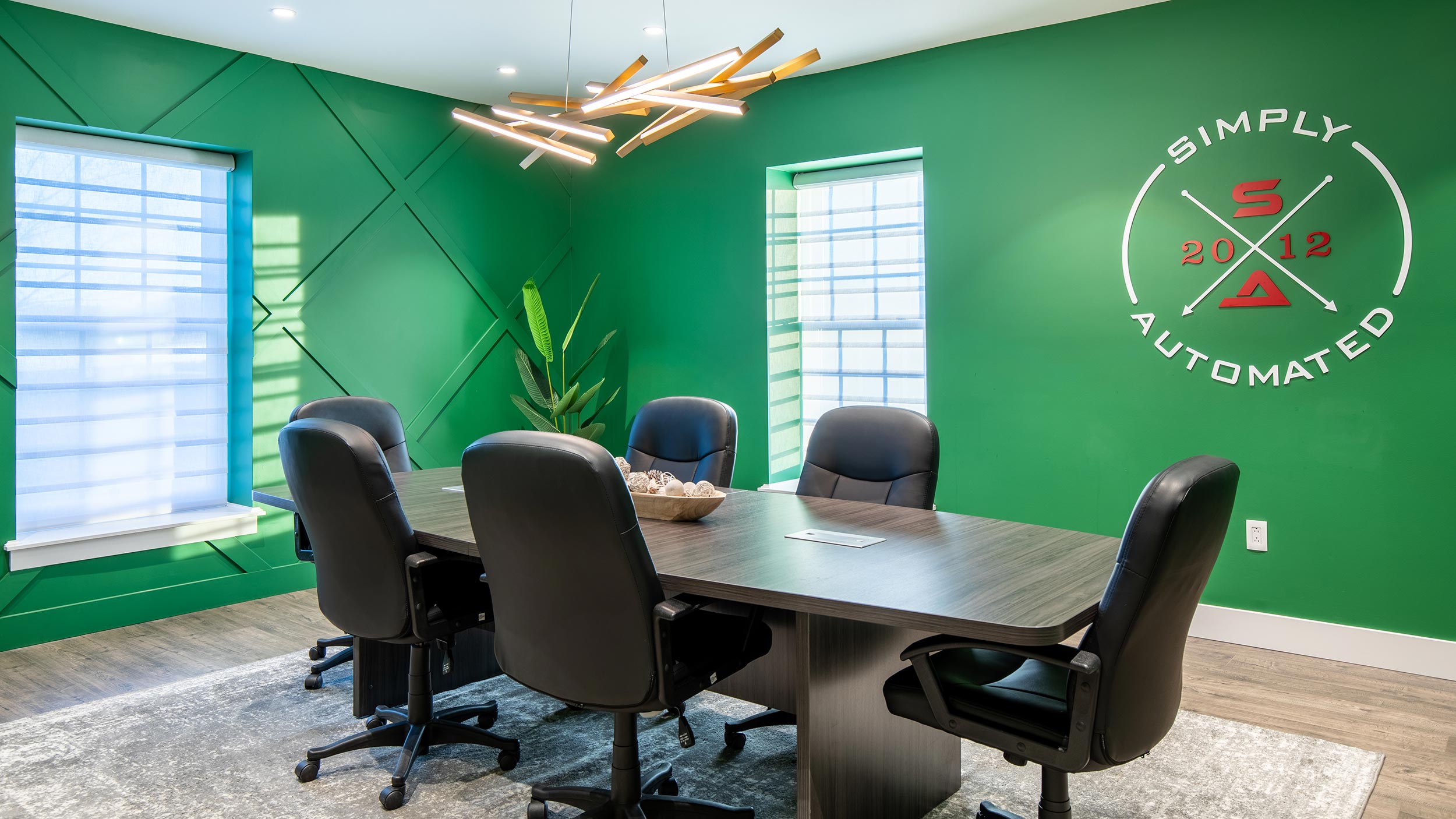Modern conference room with green walls, a wooden table, black chairs, and a unique ceiling light fixture.