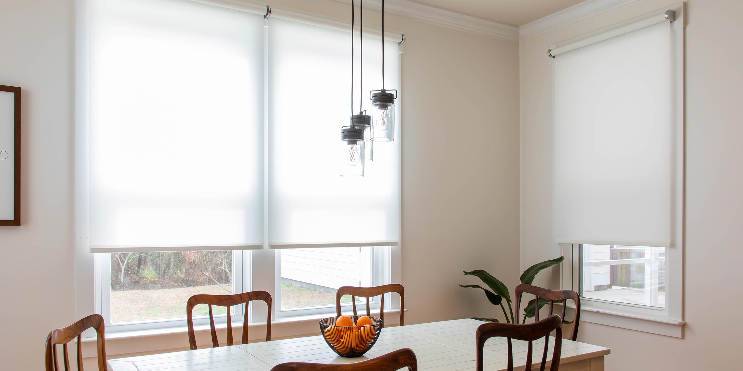 A bright dining area featuring white roller shades on large windows that softly diffuse natural light. A white table is surrounded by wooden chairs, with a bowl of oranges as a centerpiece. Three industrial-style pendant lights hang above the table, adding a modern touch to the simple, elegant space.