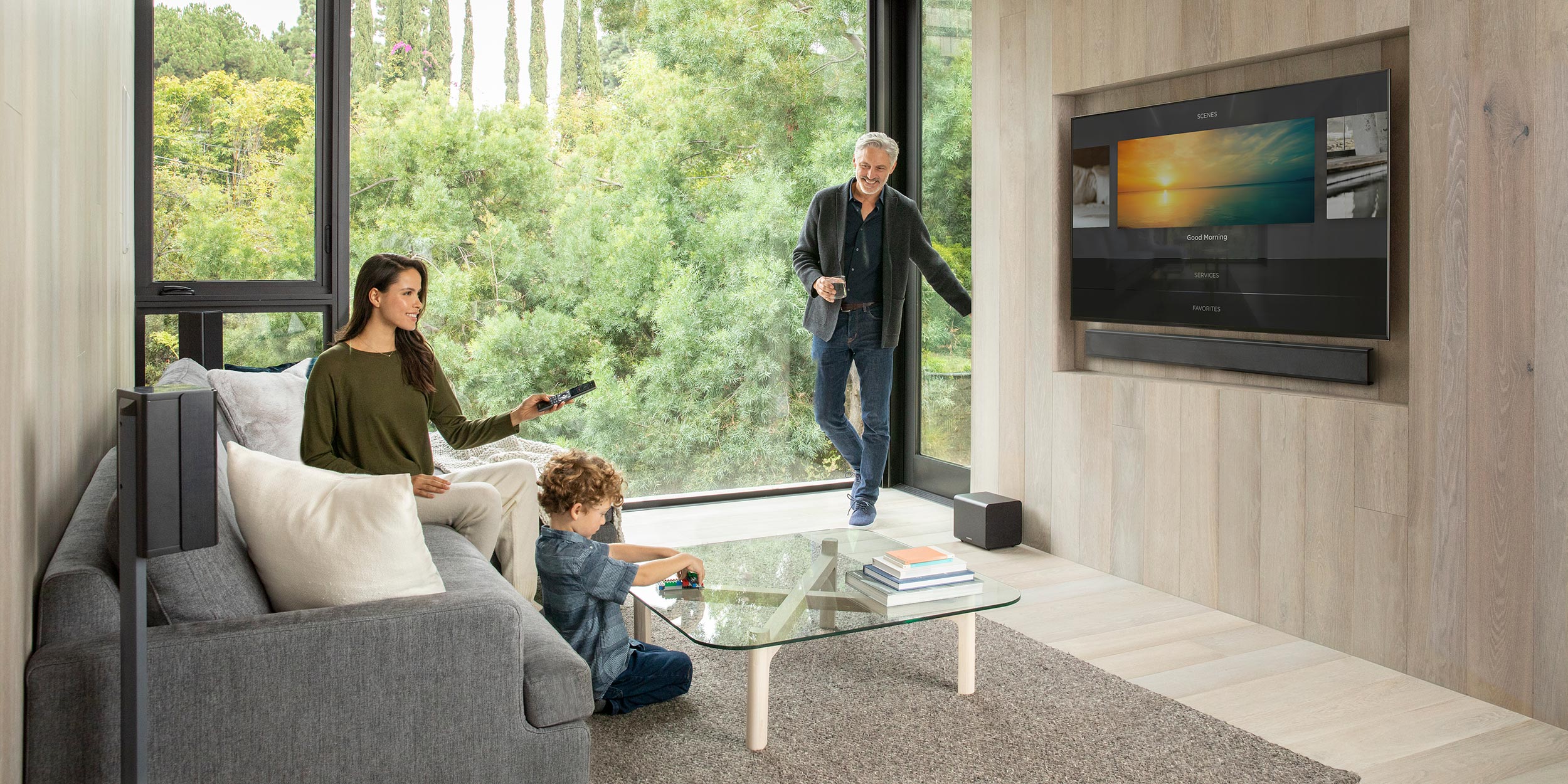 A modern living room with large windows showcasing lush greenery outside. A woman sits on a gray sofa holding a remote, a child plays with toys on a glass coffee table, and a man stands near the window with a drink, smiling. A wall-mounted TV and a sleek sound system are visible in the background, blending seamlessly into the wood-paneled wall.
